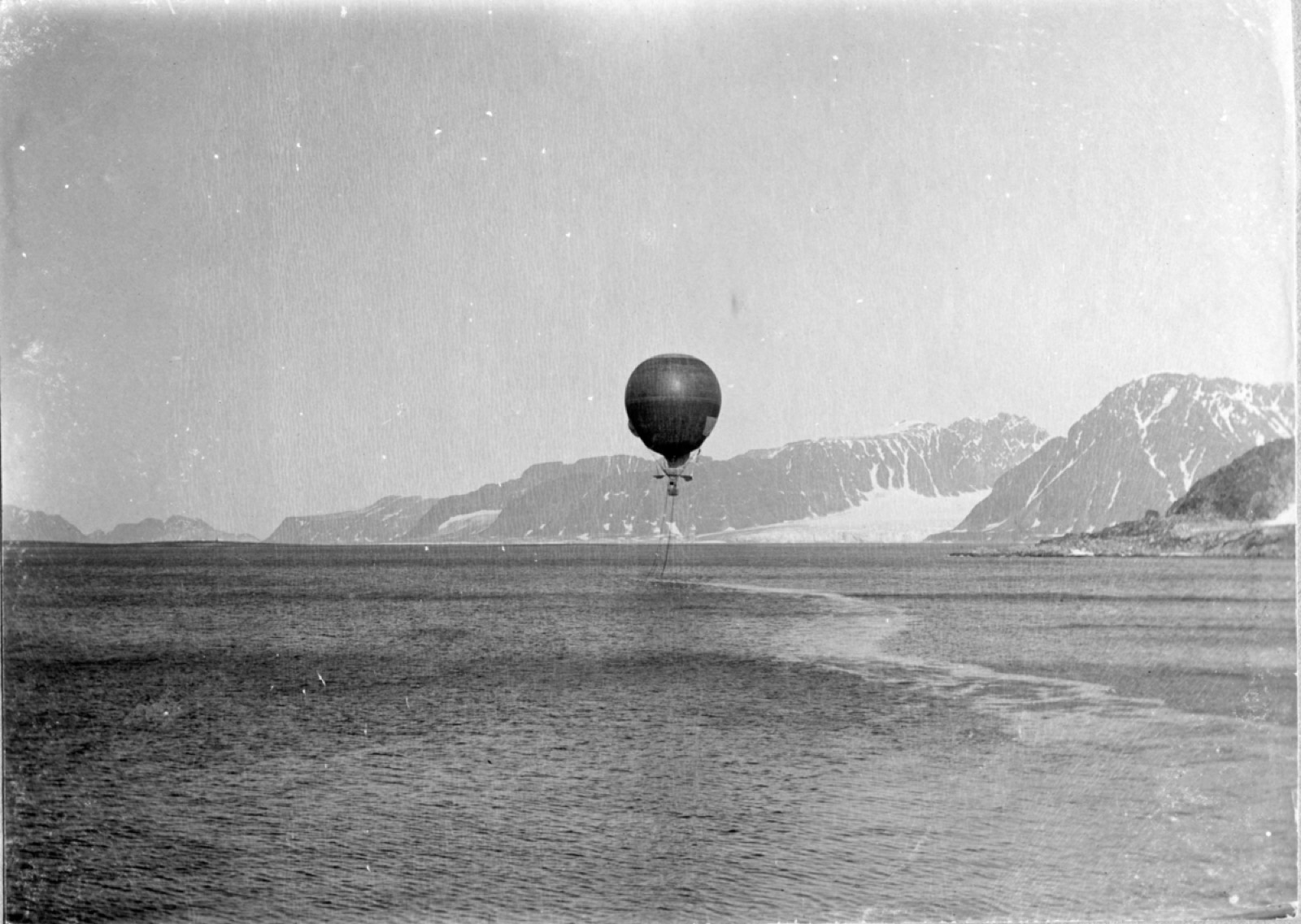 A round balloon low over a grey fjord with mountains behind, its car hanging beneath it and a wake of ropes trailing across the water