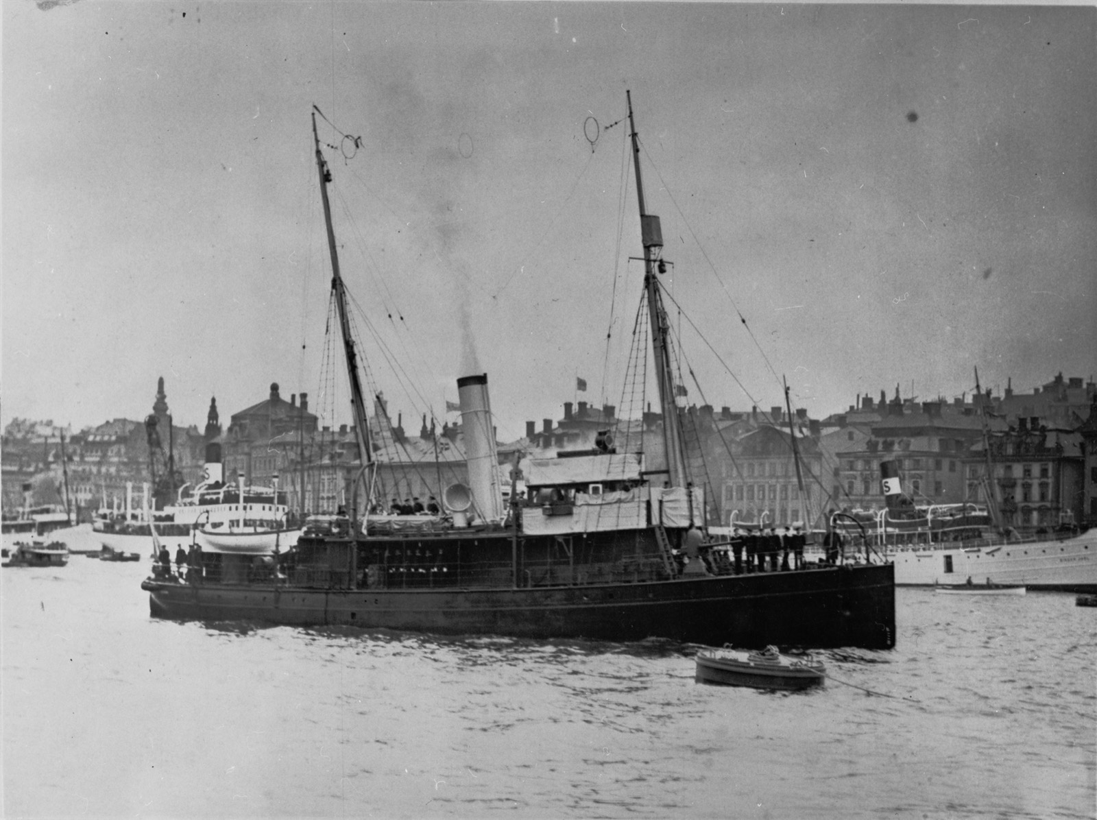 A small dark steam gunboat with two masts and one funnel on a city harbour, uniformed men along her rail, crowded quays and tall buildings behind