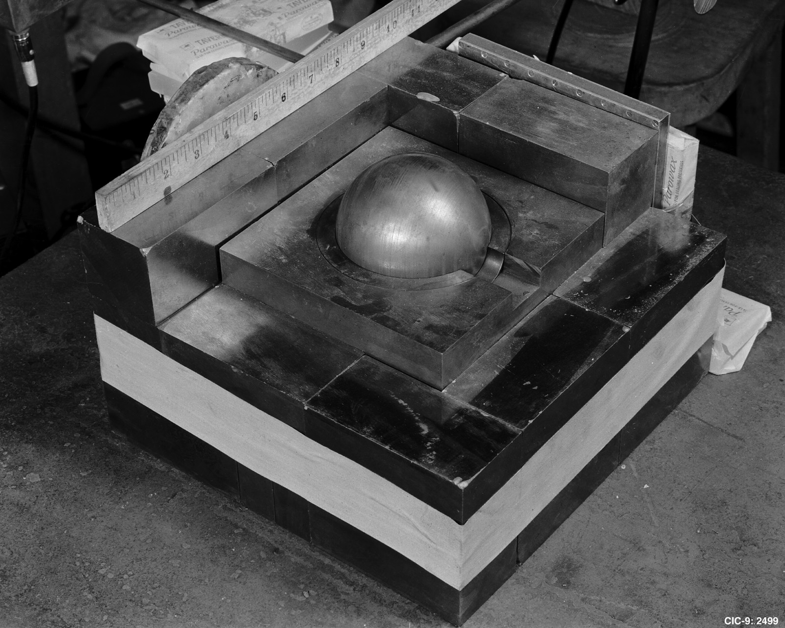 A stack of dark rectangular bricks in layers on a bench, with a polished metal hemisphere resting in a hollow at the top and a wooden ruler laid across one edge
