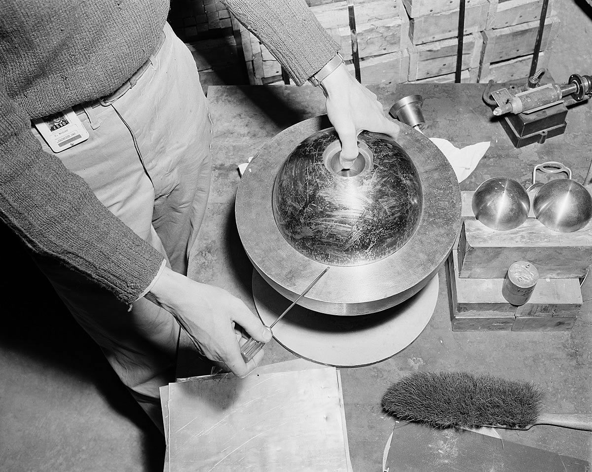 Seen from above, a man in a jumper holds a flat screwdriver under the rim of a dark metal hemisphere resting on a larger ring on a table, with his other thumb in a hole at the top of the dome; two small bright hemispheres sit on bricks beside it