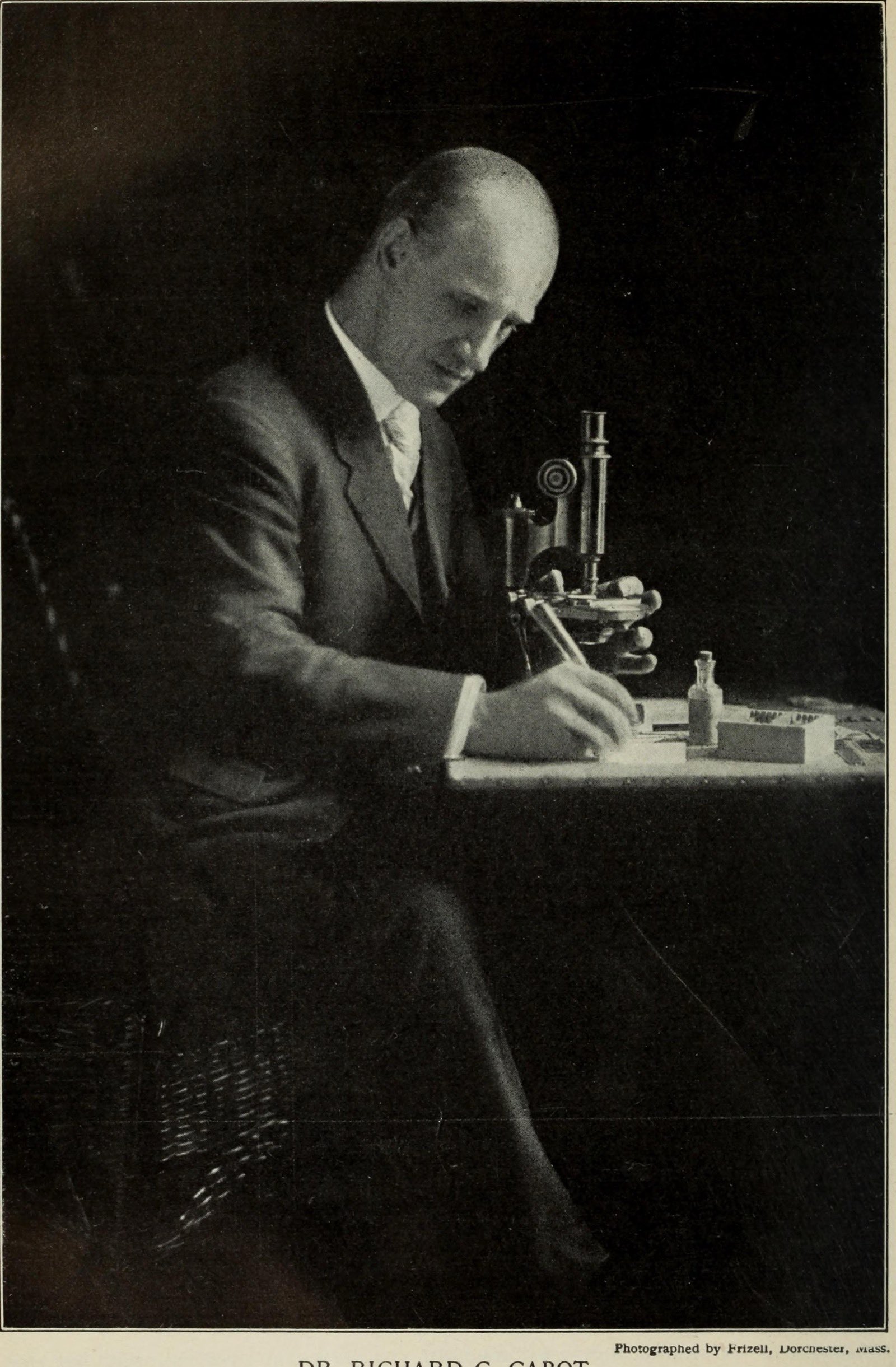 Richard Clarke Cabot, bald and in a dark suit, writing at a desk beside a brass microscope, in a dark studio photograph