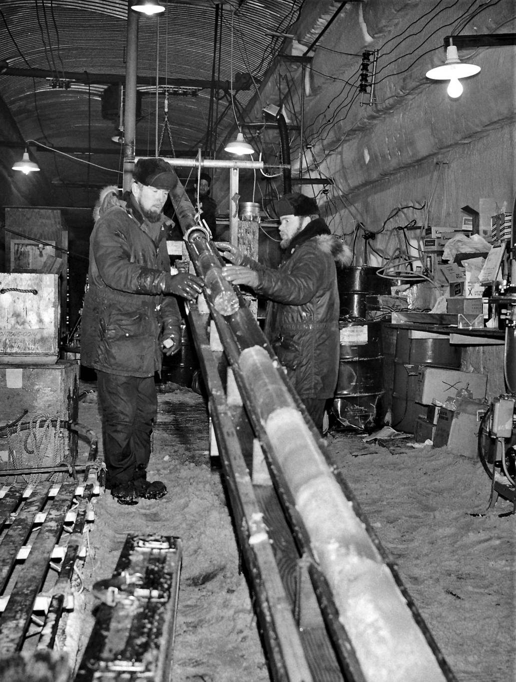 Two men in parkas inside a snow trench roofed with corrugated steel, lifting a long cylindrical ice core from its barrel onto a trough, with lamps hanging above and drums and boxes behind them