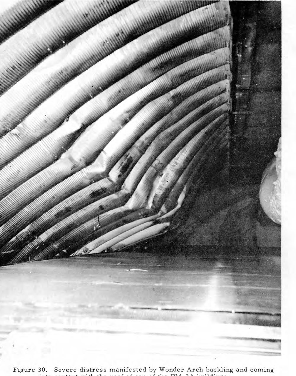 A close photograph of corrugated steel arch sections buckled and folded inward under the weight of snow, pressing down on the flat roof of a building below