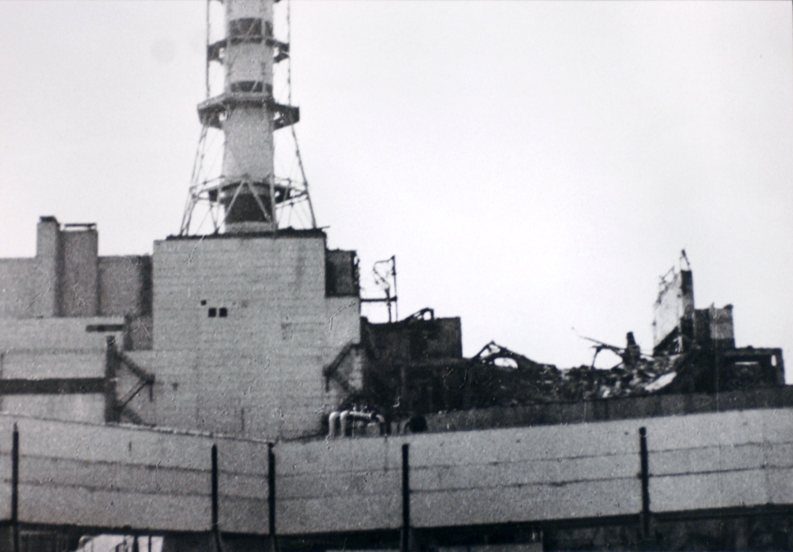 A black and white photograph of the wrecked upper storeys of the Unit 4 reactor building, with the ventilation stack behind and a long wall in the foreground