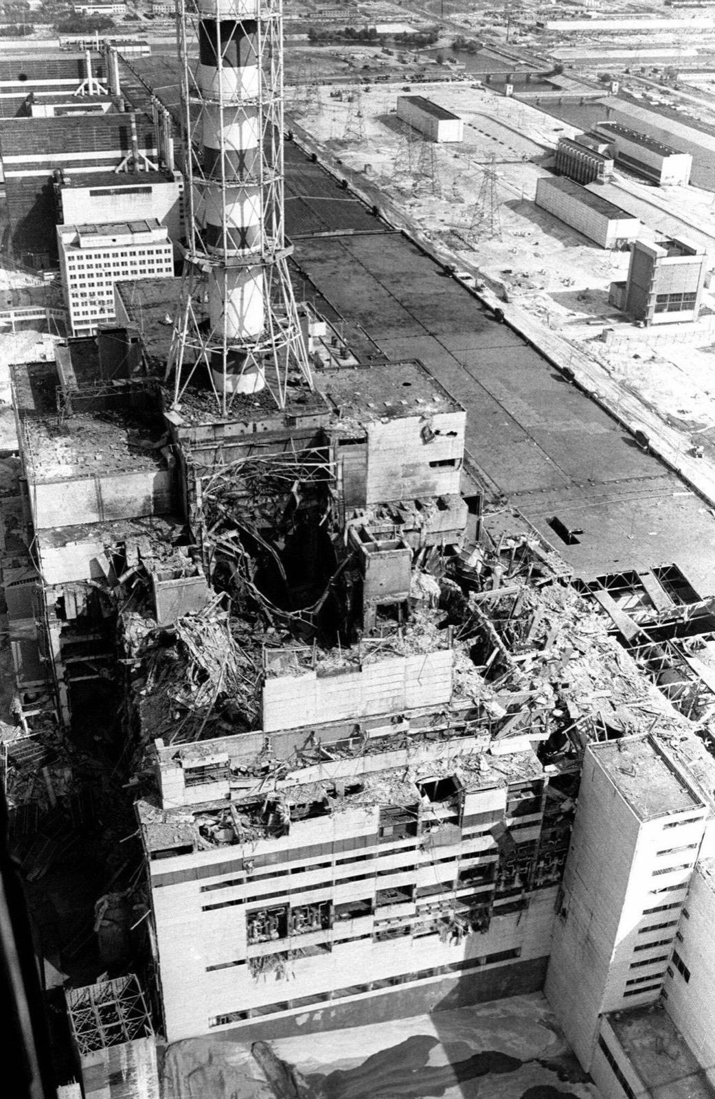 A black and white aerial photograph of the destroyed reactor building, the roof gone and the walls broken open around the ventilation stack, with the plant and the river beyond