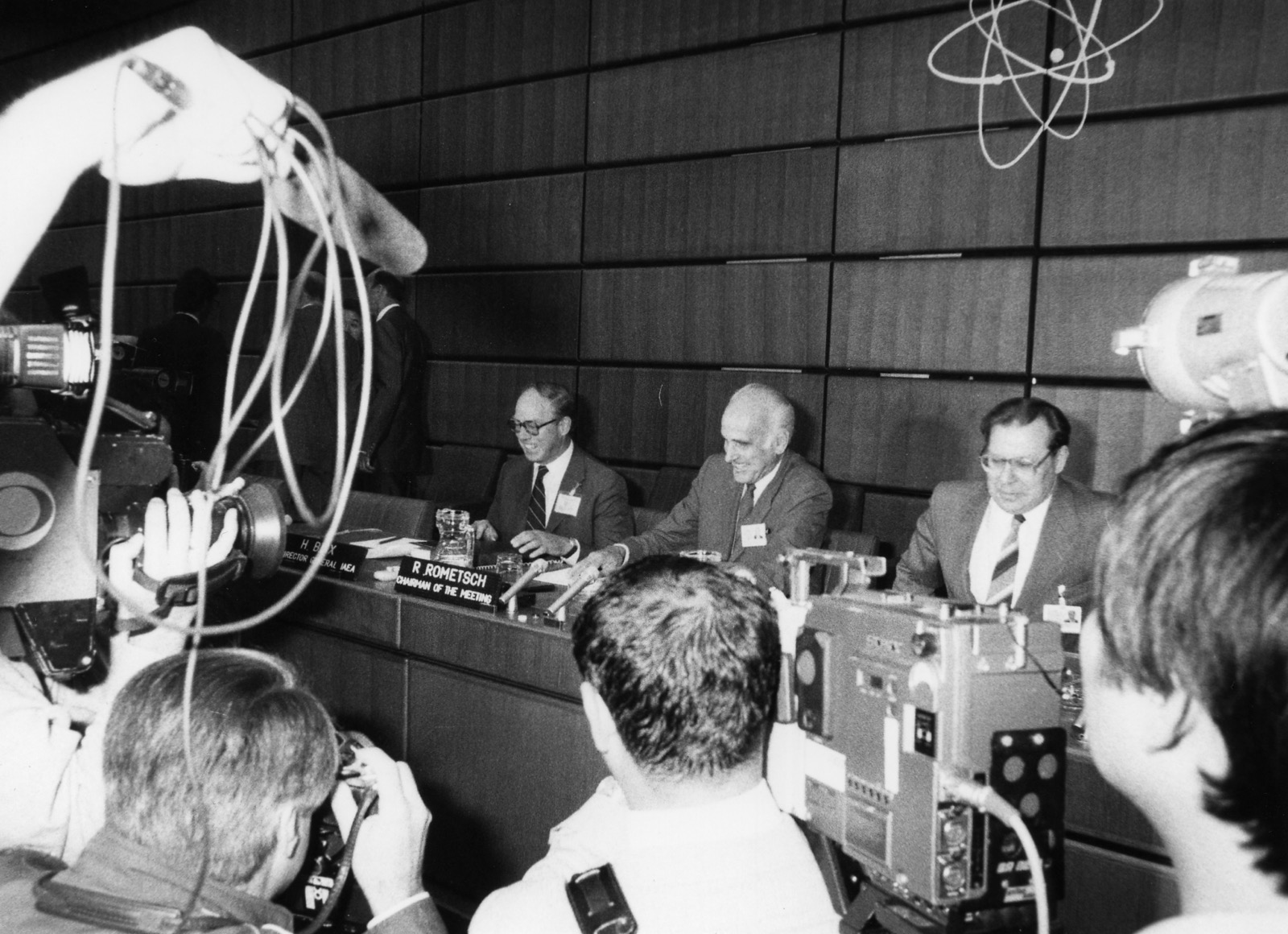 A black and white photograph of three men at a long table in a wood-panelled boardroom, with television cameras and photographers crowding in front of them