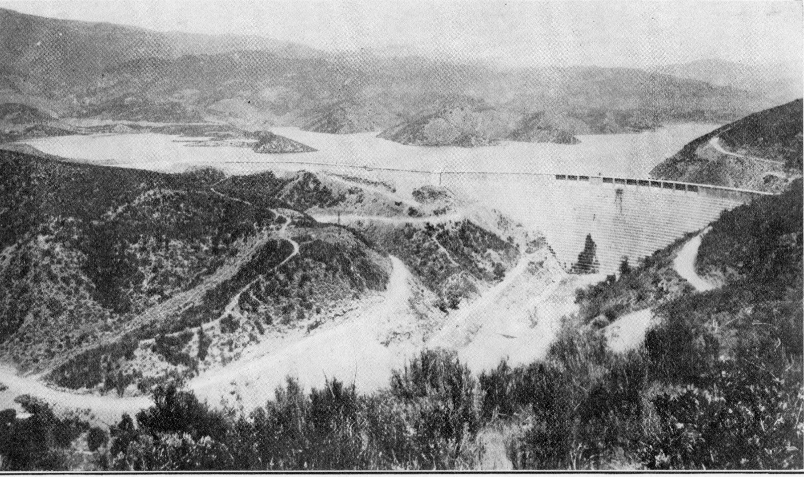 An oblique view from a hillside downstream of a stepped concrete dam curving across a canyon, with the spillway openings along its crest, a long dike running along the ridge to the right, and the full reservoir and bare hills behind