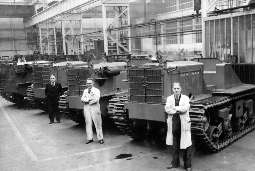 Rows of Shervick tractors, rebuilt from Sherman tank hulls, lined up inside a large factory shop with workmen beside them