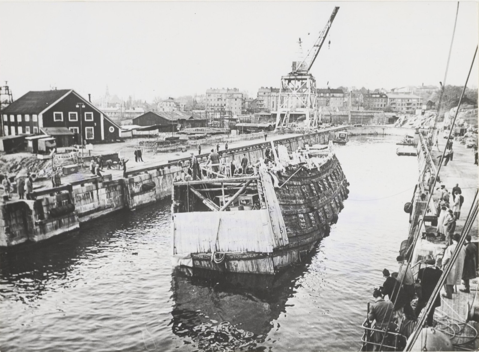 The raised hull of the Vasa, dark and encrusted, floating on her own keel between stone quay walls as she is moved into a dry dock, with workers on her deck and spectators along the quay