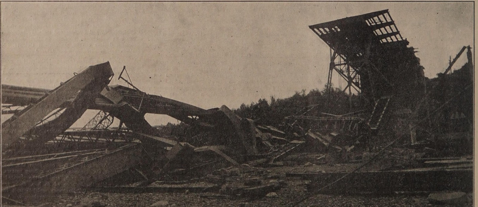 Long bent steel chords and trusses of the fallen anchor arm lying along the ground, with the tall latticed frame of the erection traveller tilted over at the right and trees behind