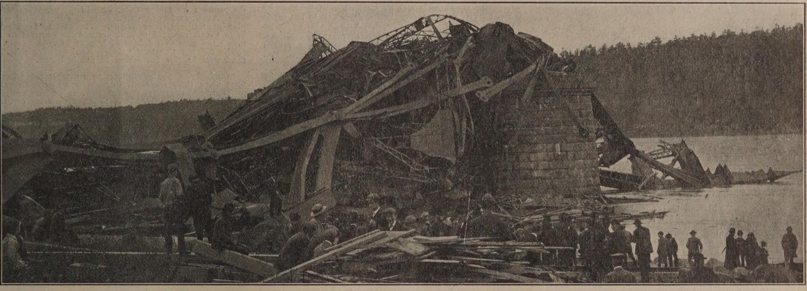 A tangle of enormous bent steel members heaped against and over a tall stone pier at the water's edge, with a crowd of men in hats standing on the wreckage and along the shore, and a wooded bank across the river