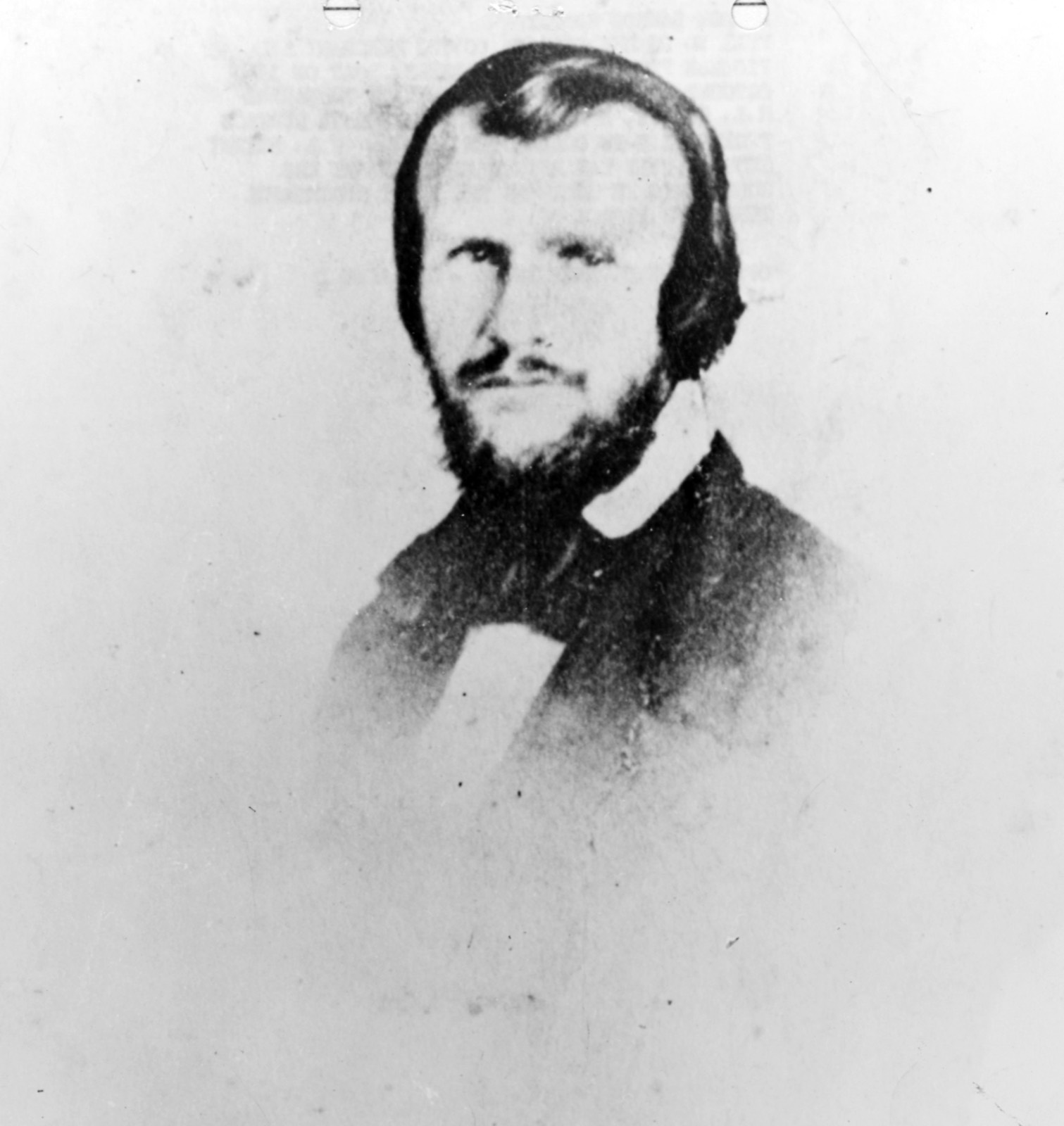 A studio portrait of a bearded man in a dark coat and white collar, faded, with his hair combed back