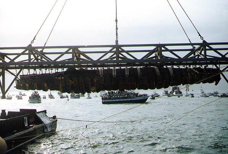 A long dark encrusted hull hanging in a row of slings beneath a steel truss above the sea, with a crowd of boats watching behind it