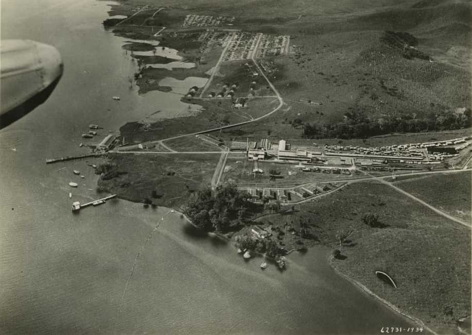 An aerial photograph of Fordlandia in 1934: the sawmill and dock on the river bank, rows of workers' houses behind, and cleared hills beyond